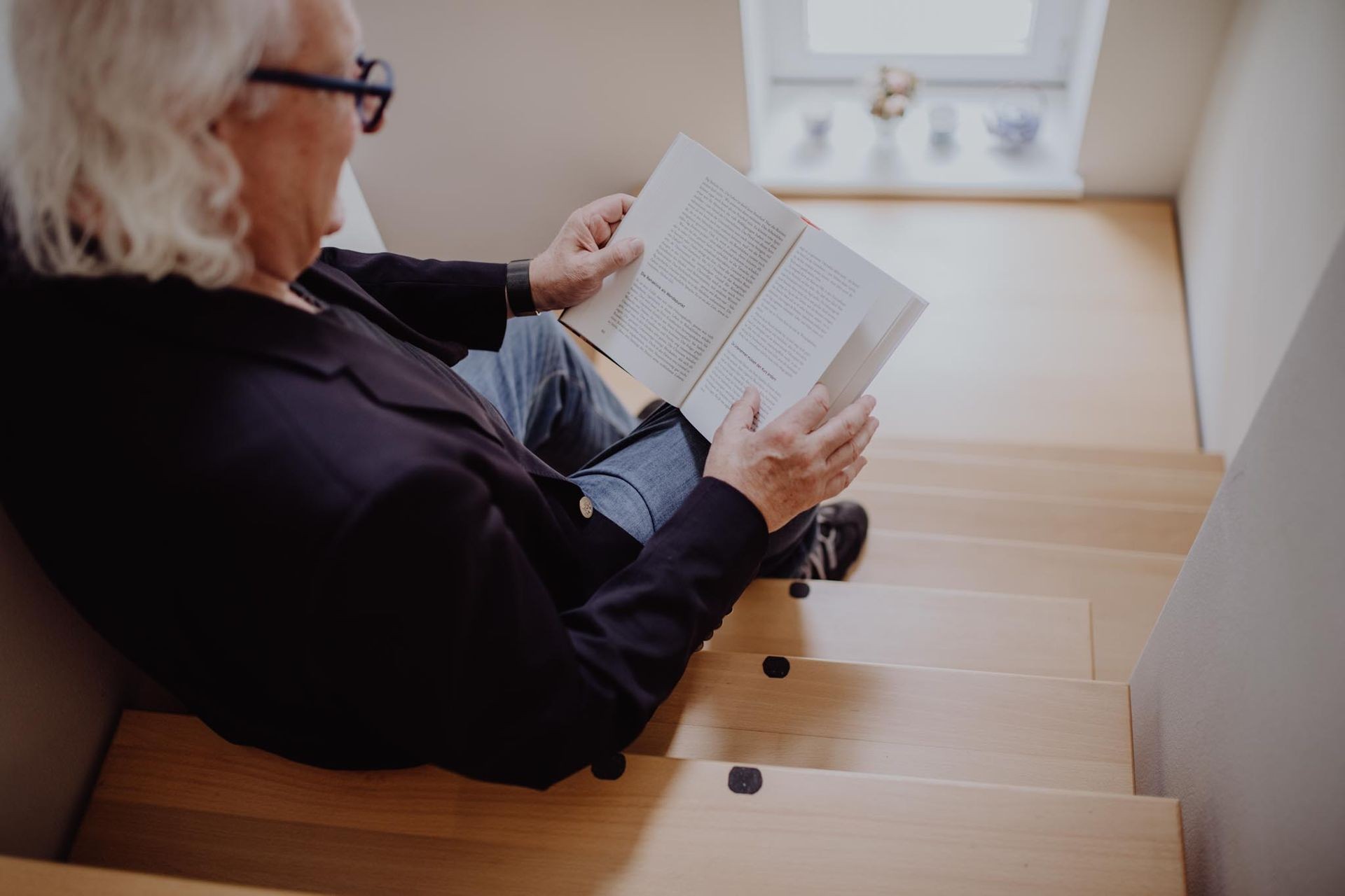 Person sitting on stairs reading a book near a window.