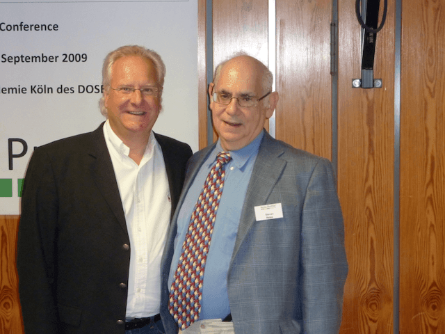 Two men standing together at a conference, wooden wall and conference sign in the background.